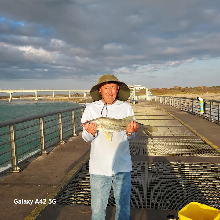 A man wearing a hat holds a fish on a pier with water and a cloudy sky in the background.