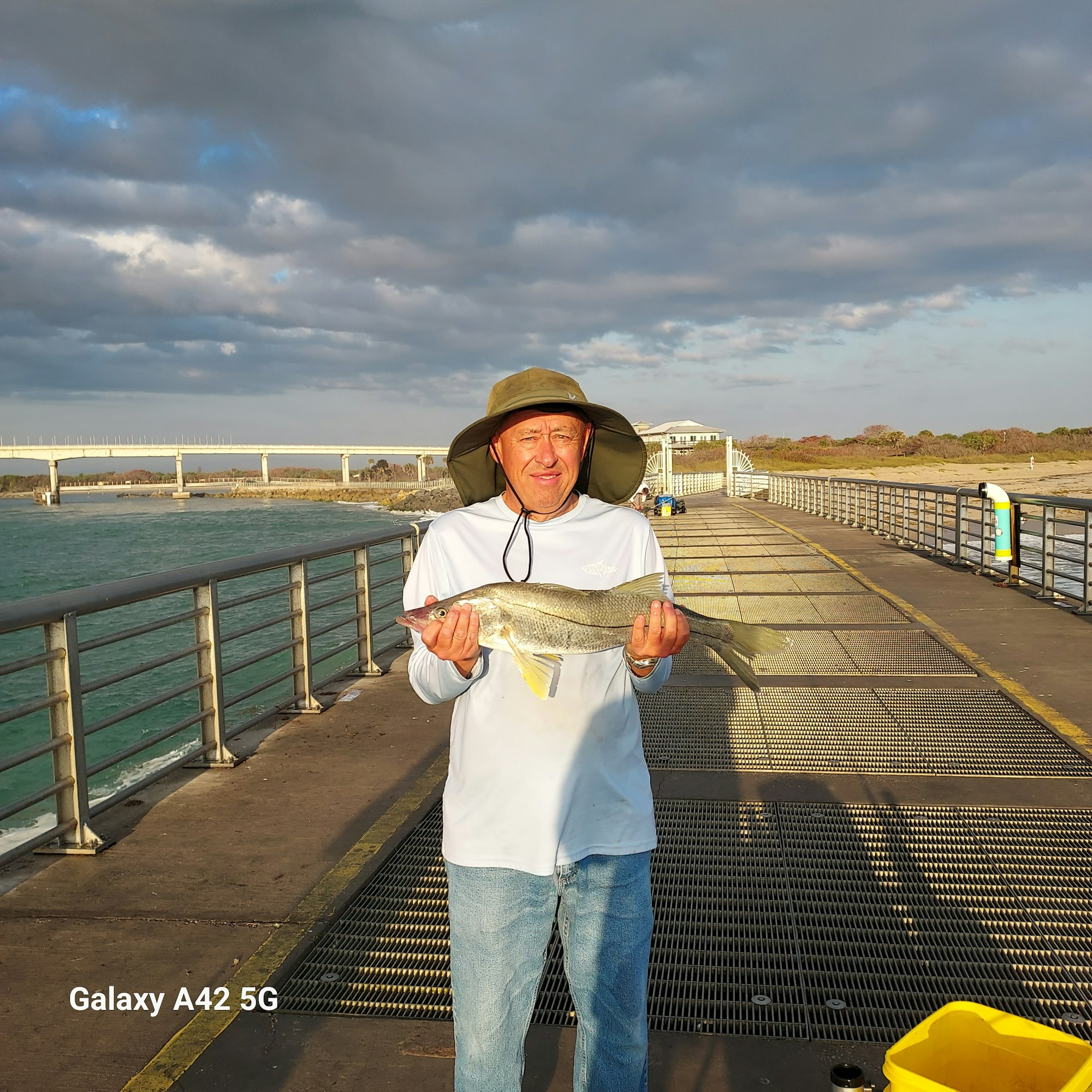 A man wearing a hat holds a fish on a pier with water and a cloudy sky in the background.