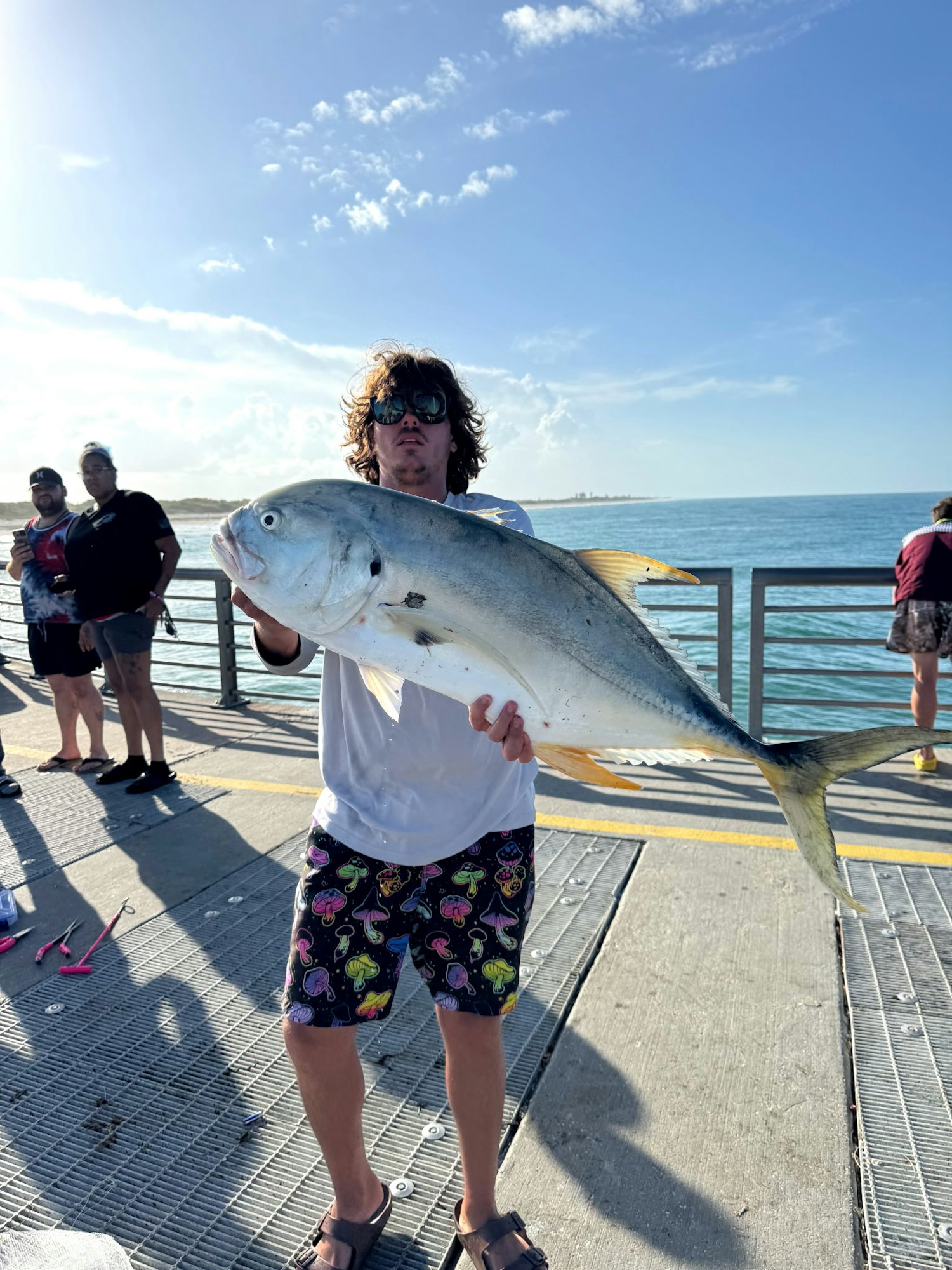 A person holding a large fish on a pier with the ocean in the background, surrounded by a few other people.