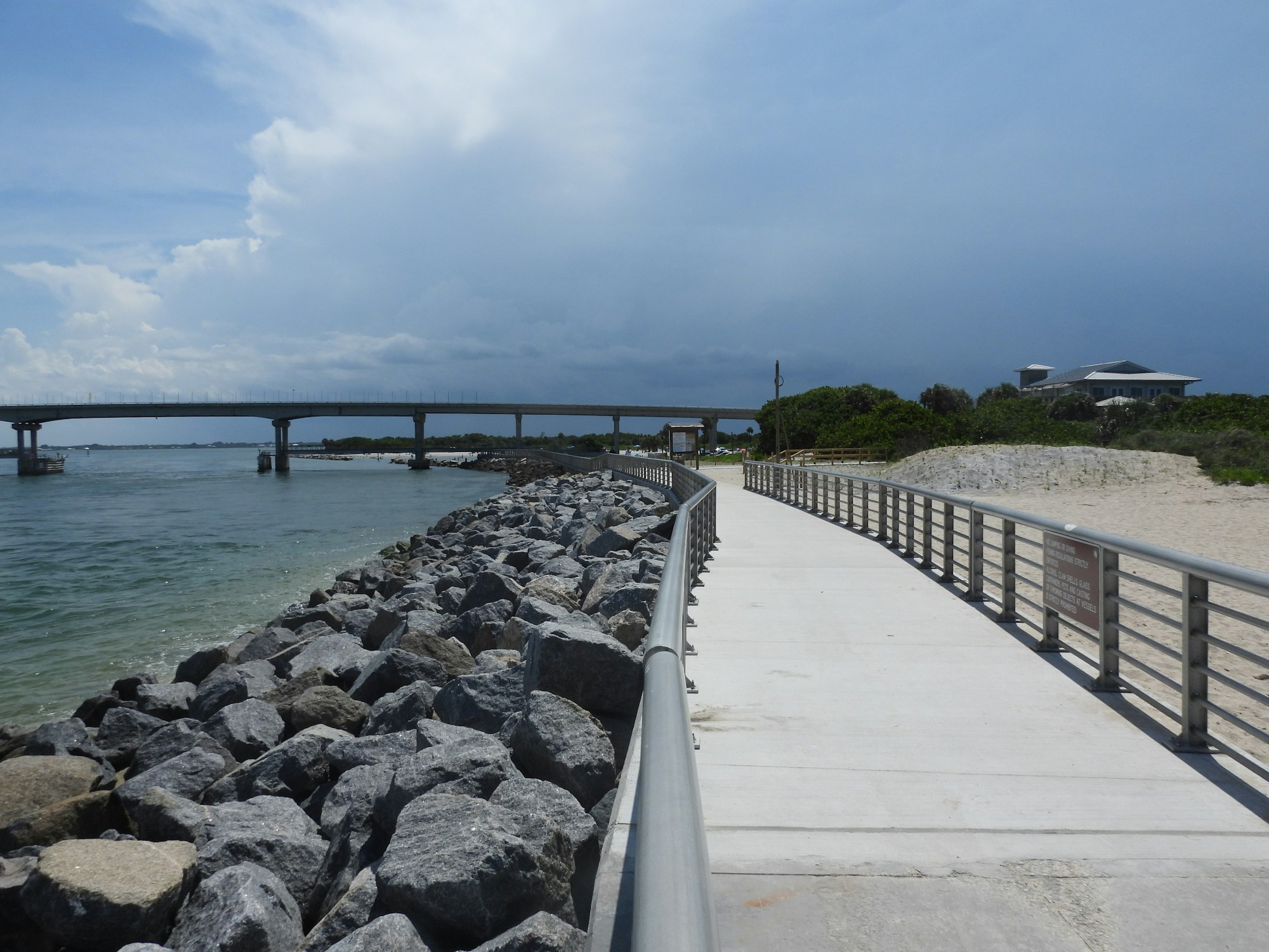 Pathway with railing alongside rocks and water, bridge in the background, cloudy sky.