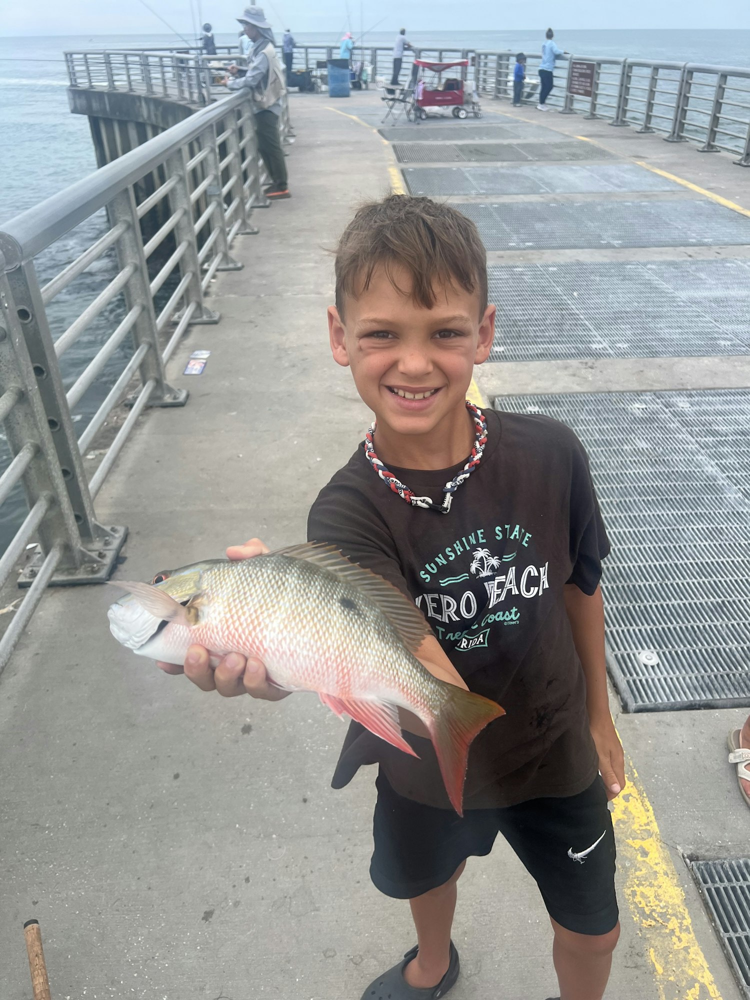 A boy smiling while holding a fish with a pier in the background. The photo is rotated 90 degrees counter-clockwise.