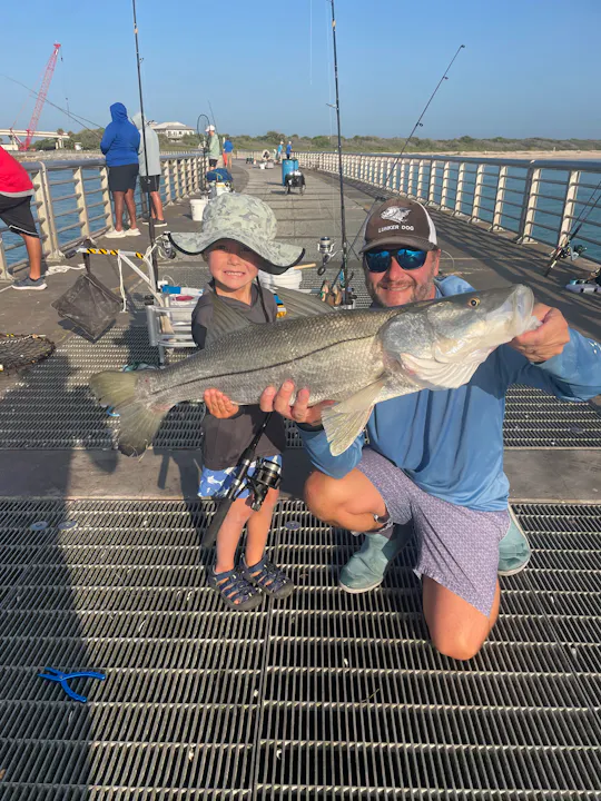 A person holding a large fish, smiling with a child nearby, on a fishing pier with other fishermen in the background.
