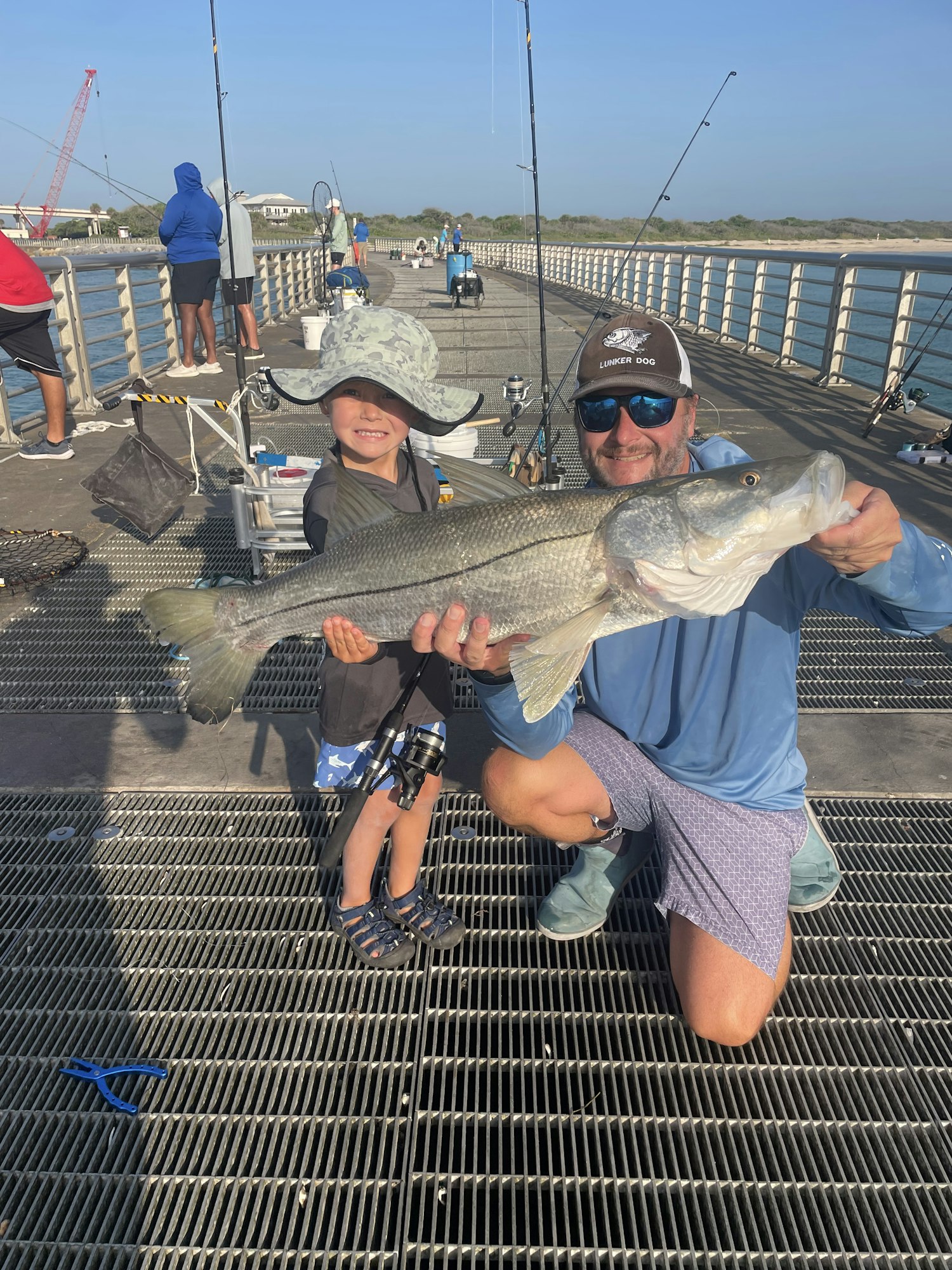 A person holding a large fish, smiling with a child nearby, on a fishing pier with other fishermen in the background.