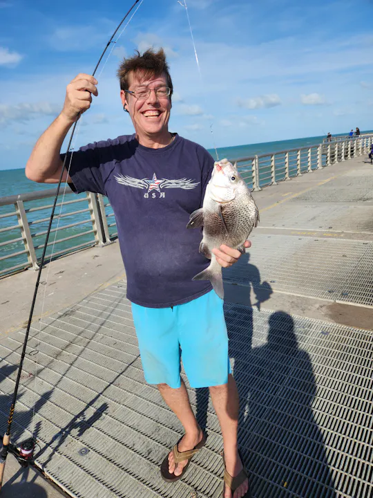 A smiling person in shorts holds a fish on a pier with the ocean in the background, enjoying a day of fishing.