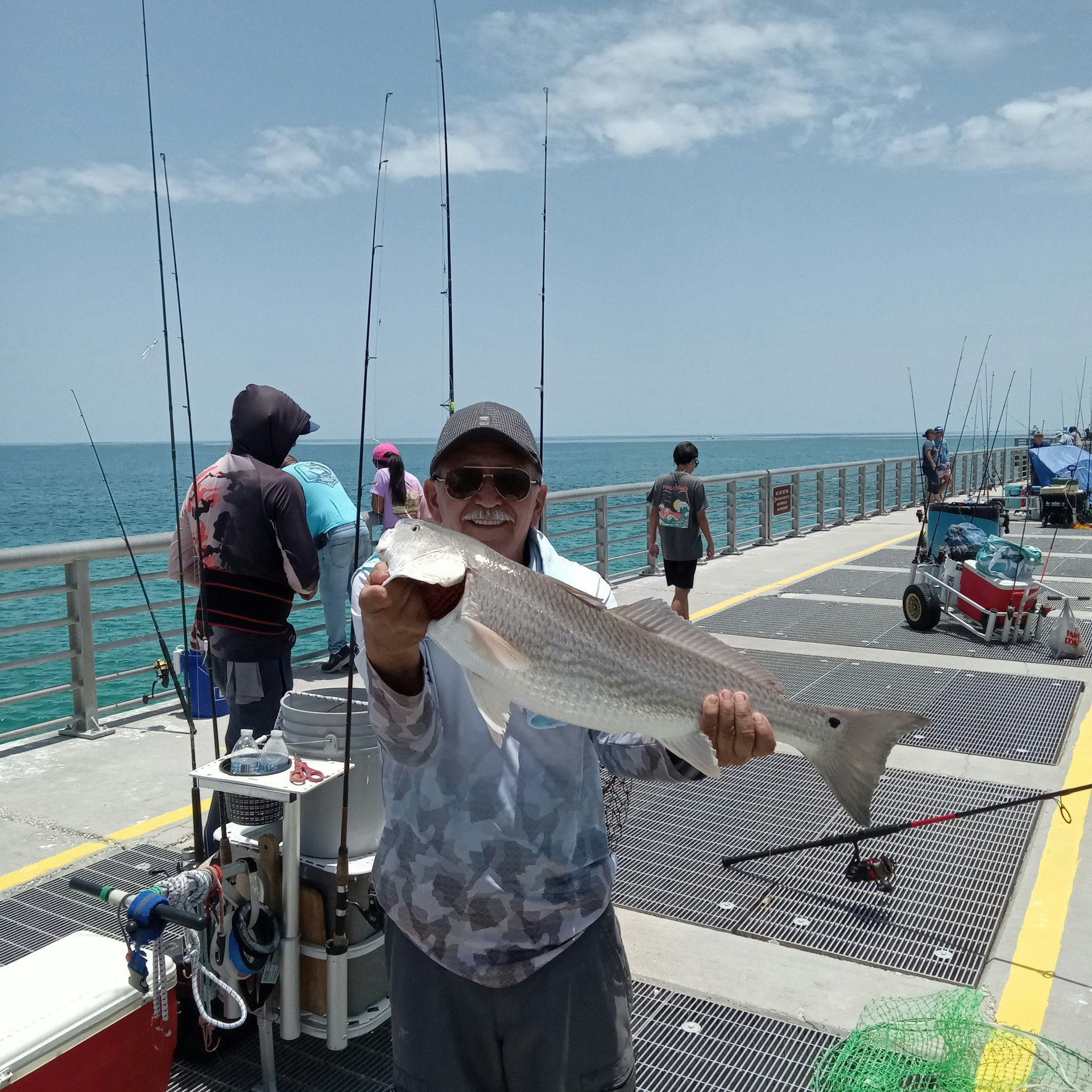 A person holding a large fish on a pier, surrounded by fishing gear and other people, with the ocean in the background.