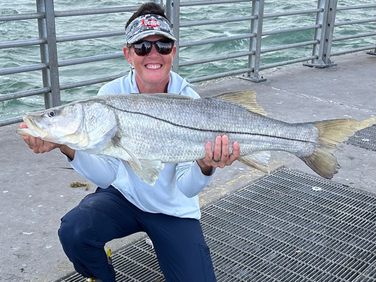 A person holding a large fish at a waterfront location with a railing in the background.