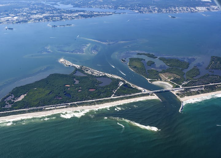 Aerial view of a coastline with inlets, islands, a bridge, boats, and waves breaking on a sandy beach.