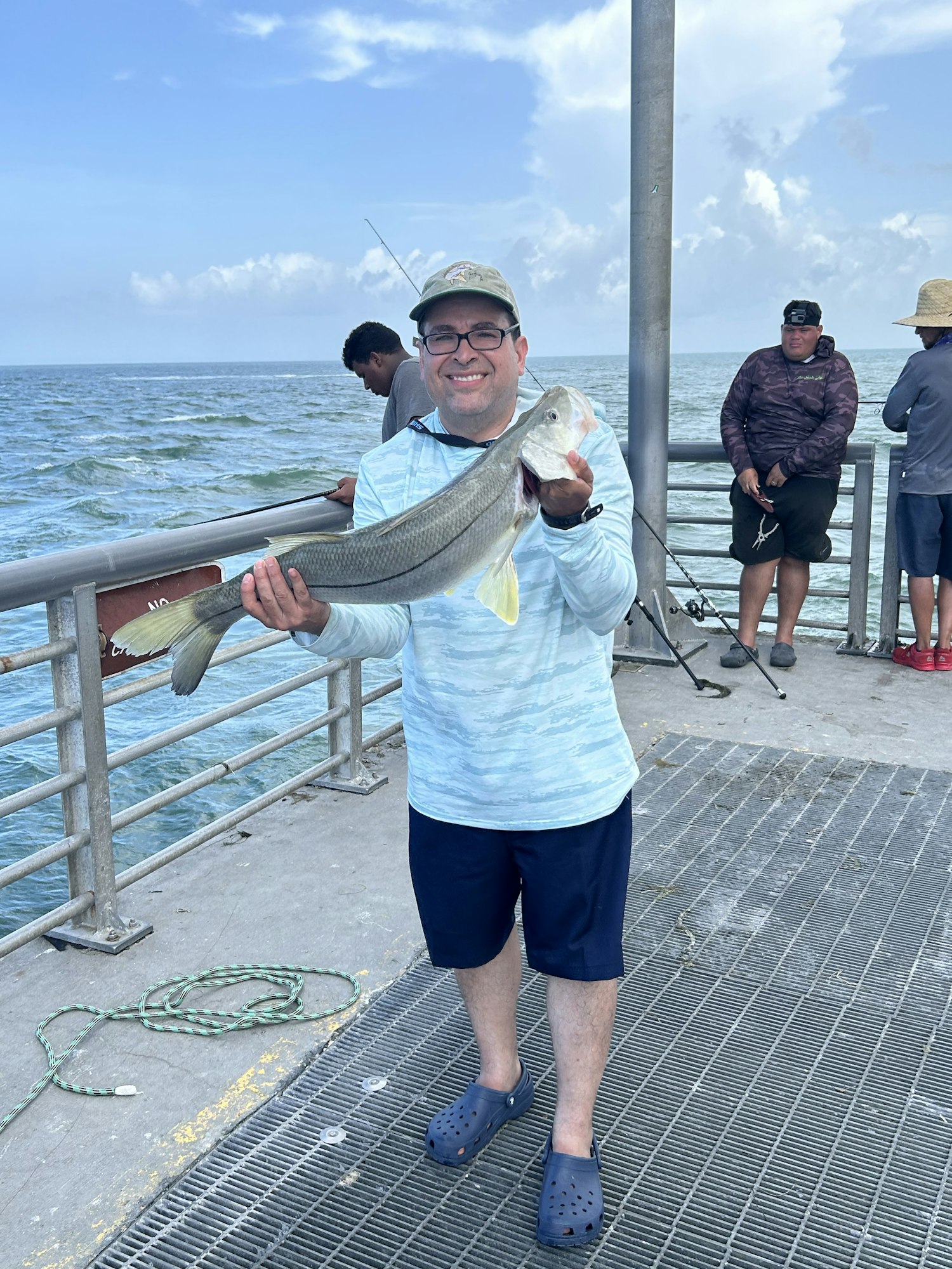 A person holding a fish on a pier, with others fishing in the background. Image is rotated 90 degrees.
