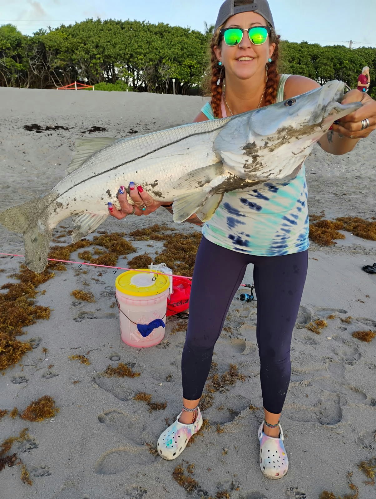Person holding a large fish on a beach, wearing sunglasses and casual attire.
