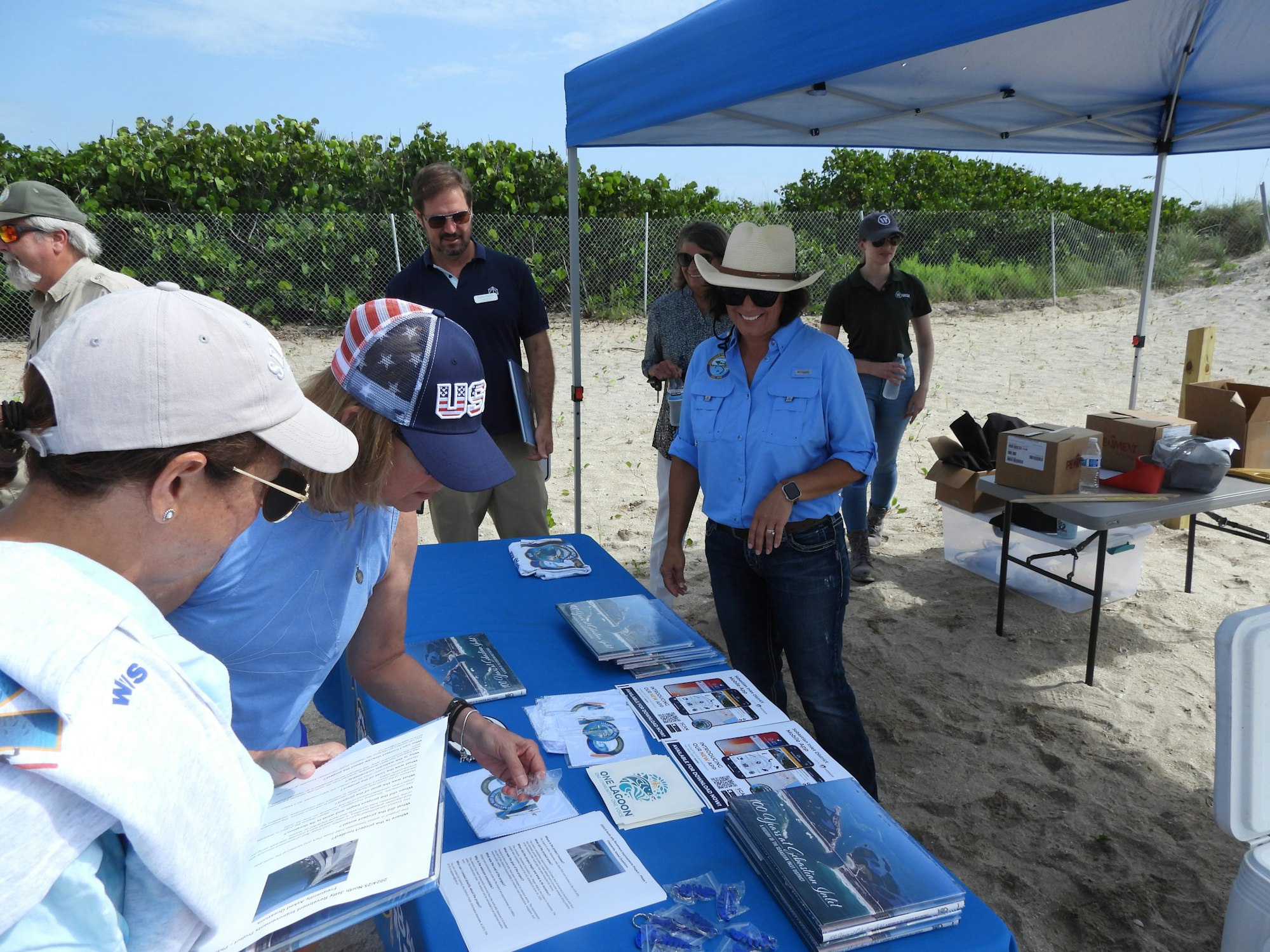 People at an outdoor booth with brochures and flyers under a canopy on a sandy beach.