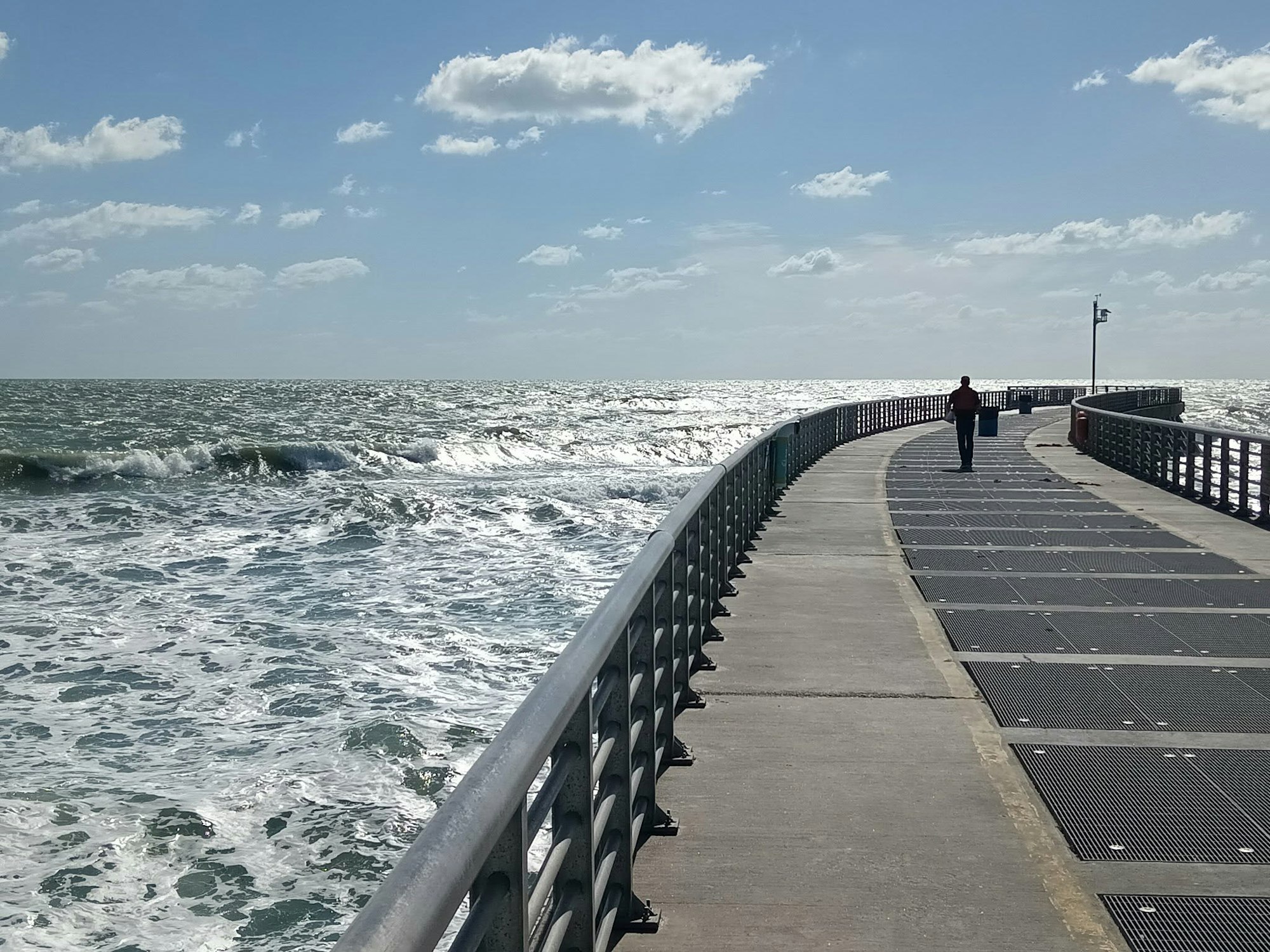 A person walks along a curved pier, with waves crashing against it, under a bright blue sky with fluffy clouds.