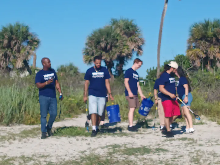 A group of people in matching blue shirts engaging in beach clean-up activities, carrying buckets and tools on a sunny day.