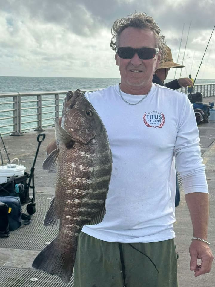 A man wearing sunglasses holds a large fish while fishing on a pier, with fishing equipment visible in the background.