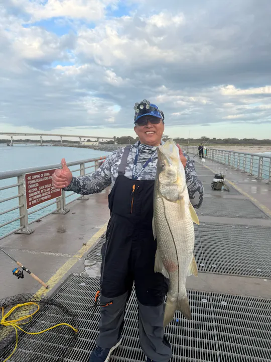 A person on a pier proudly holds a large fish, smiling, with fishing gear and a scenic water backdrop under cloudy skies.