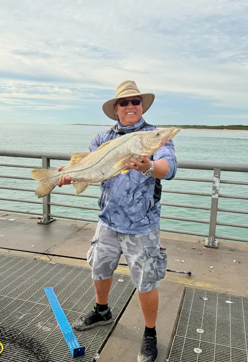 A person on a pier proudly holds a large fish, wearing sunglasses and a hat, with water in the background.