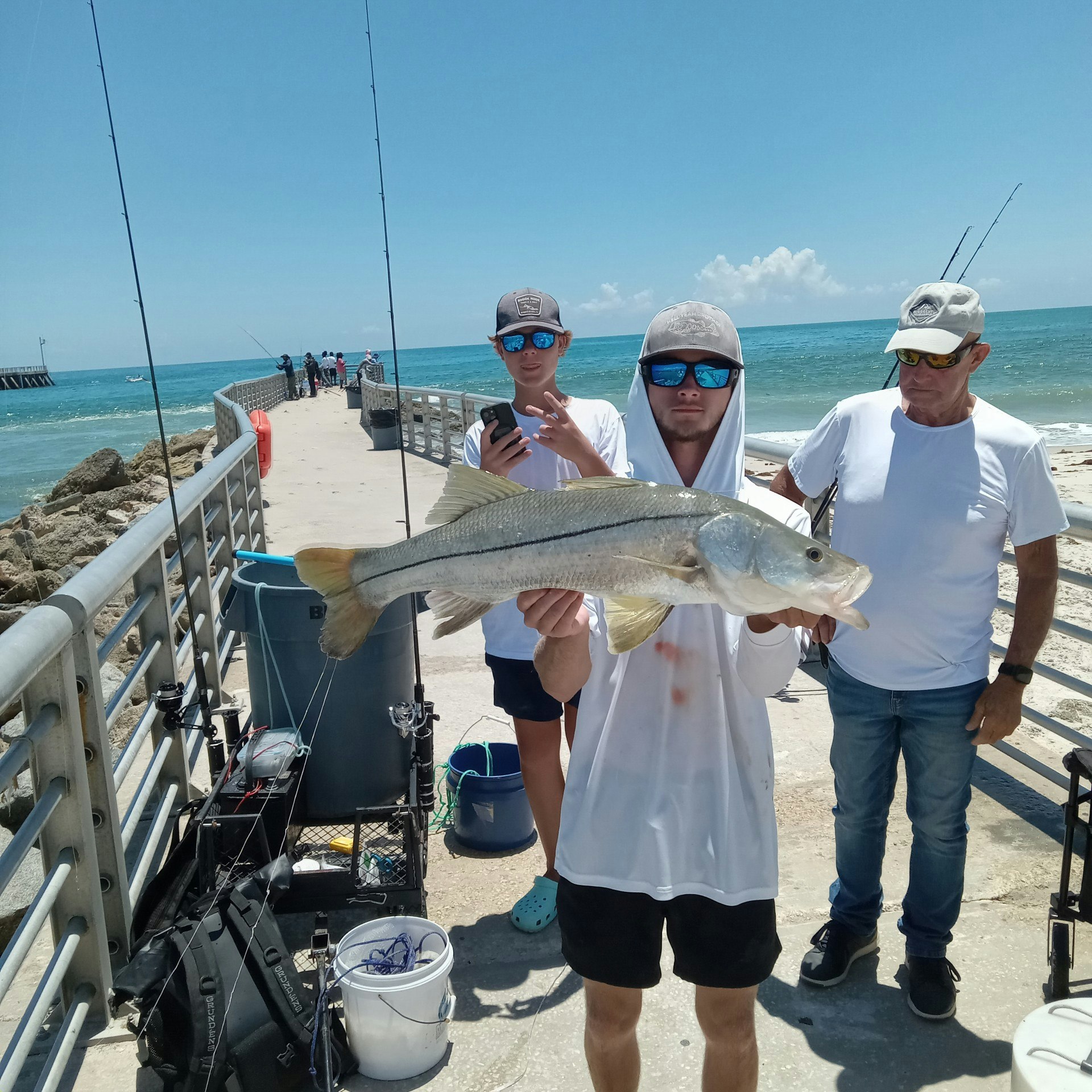 Three people on a pier by the ocean, with one holding a large fish. Two others are watching, with fishing gear around them.