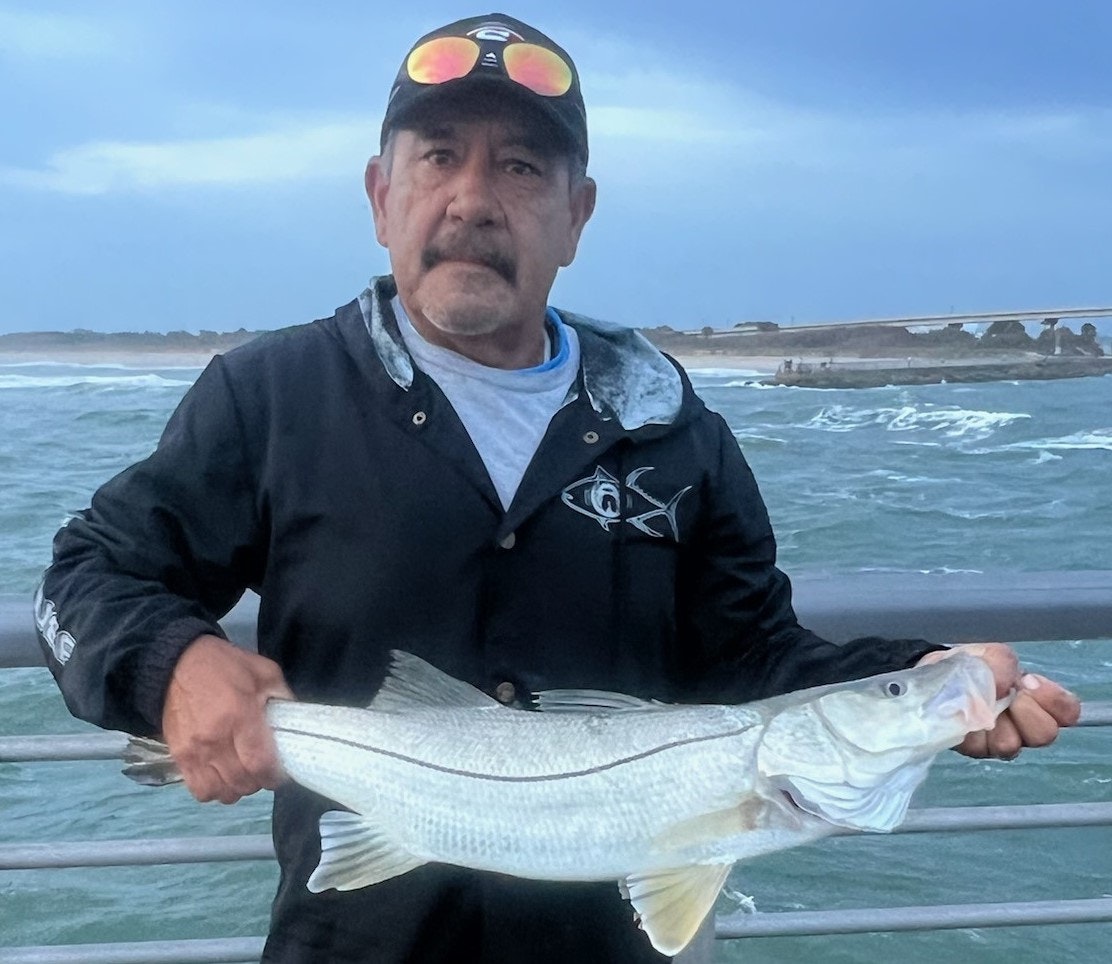 A man holding a fish on a pier with rough seas and cloudy sky in the background.