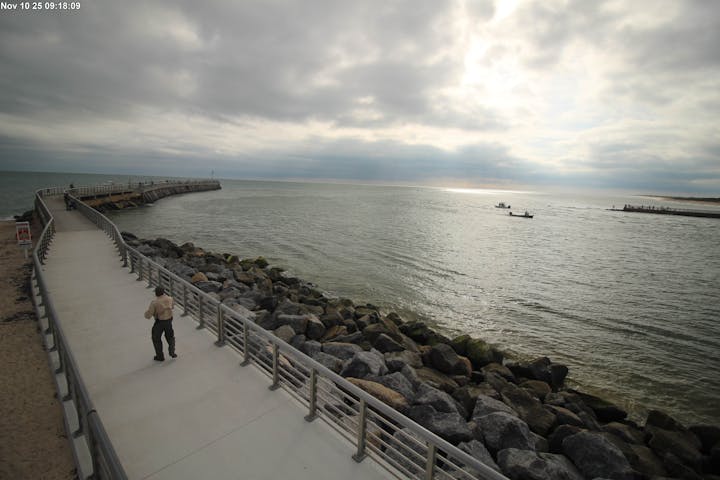 A boardwalk by a rocky shoreline with a person walking, cloudy sky, and boats in the distance.
