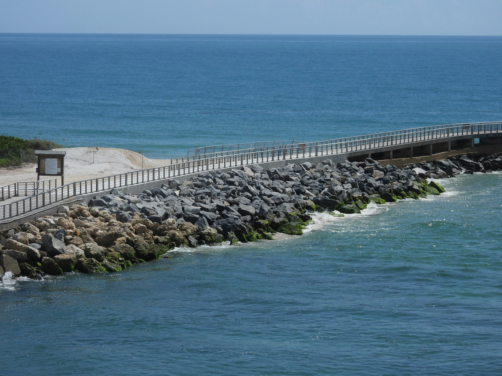 Rocky jetty with railing extends into the ocean, waves crashing, clear blue water and sky, sandy walkway on the left.