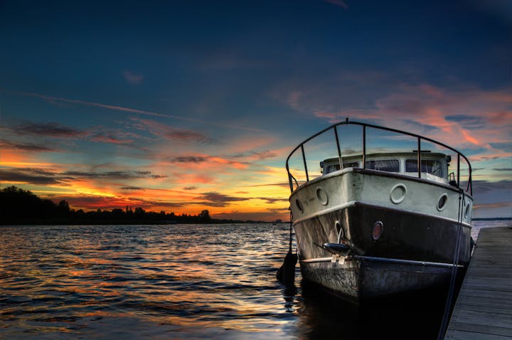 A boat rests by the shore, reflecting sunset colors on the water, with a vibrant sky filled with clouds.