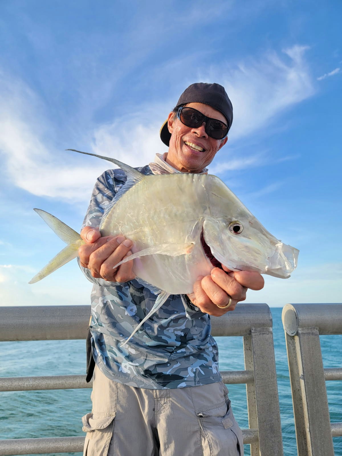 A person in a cap and sunglasses holding a fish on a pier, with a blue sky and ocean in the background.