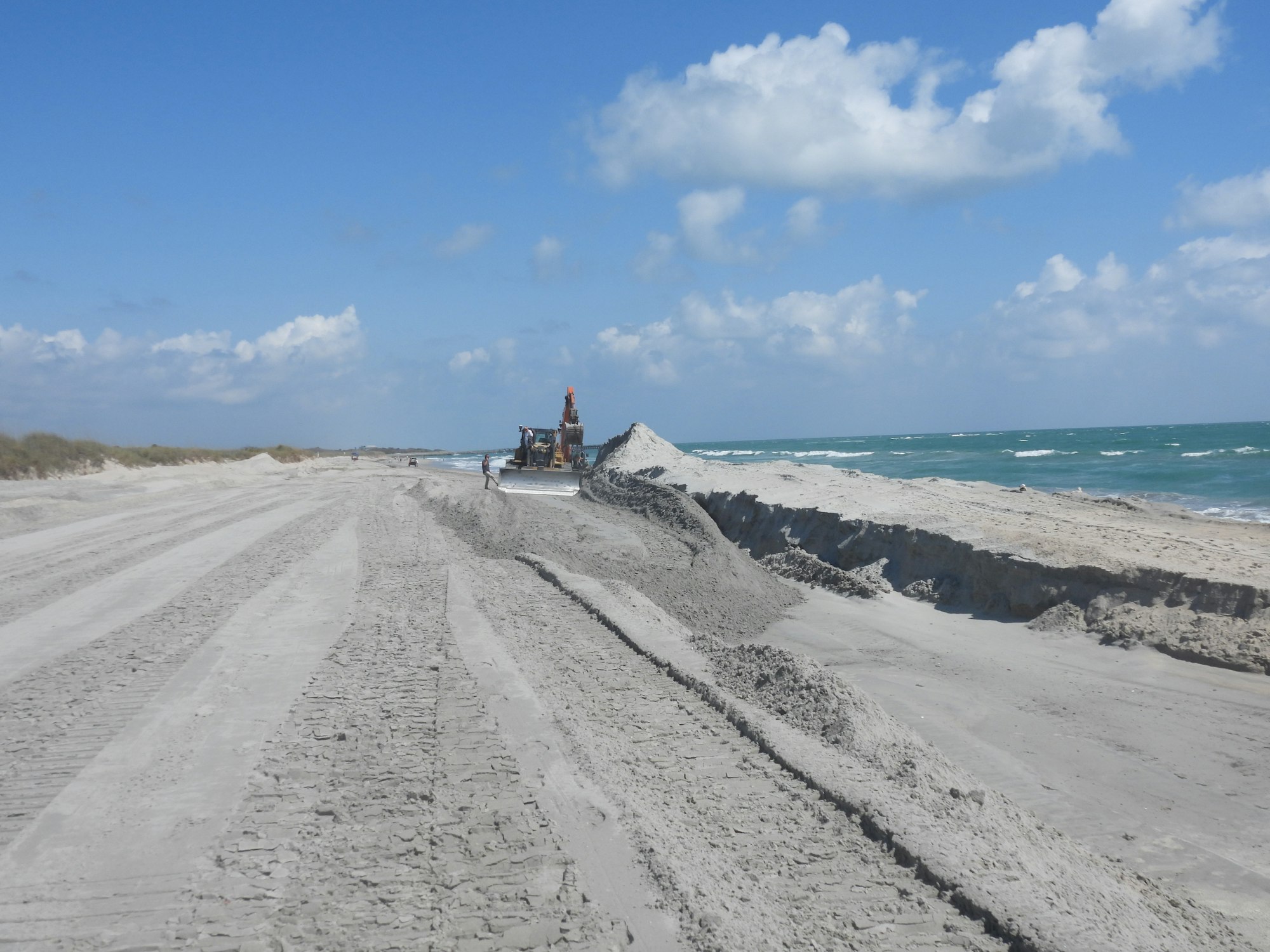 A beach scene with machinery, tire tracks in sand, and waves under a blue sky with clouds, suggesting maintenance or construction work.