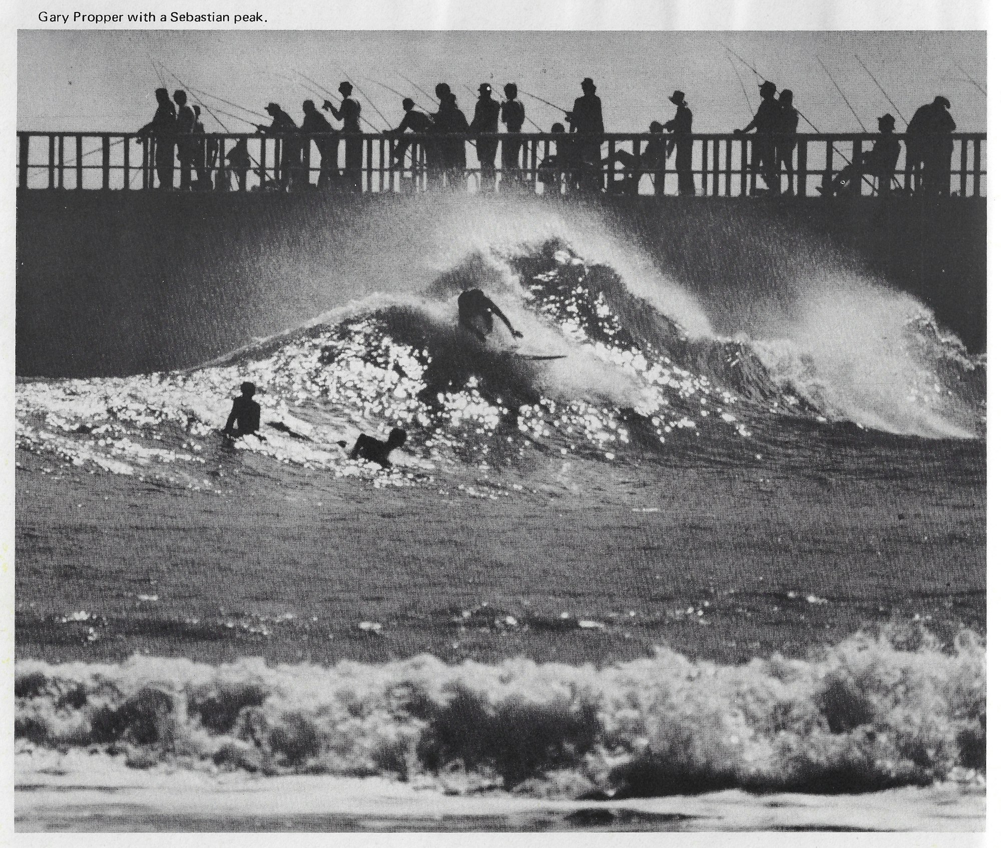 surfers riding a wave with fisherman on pier in background
