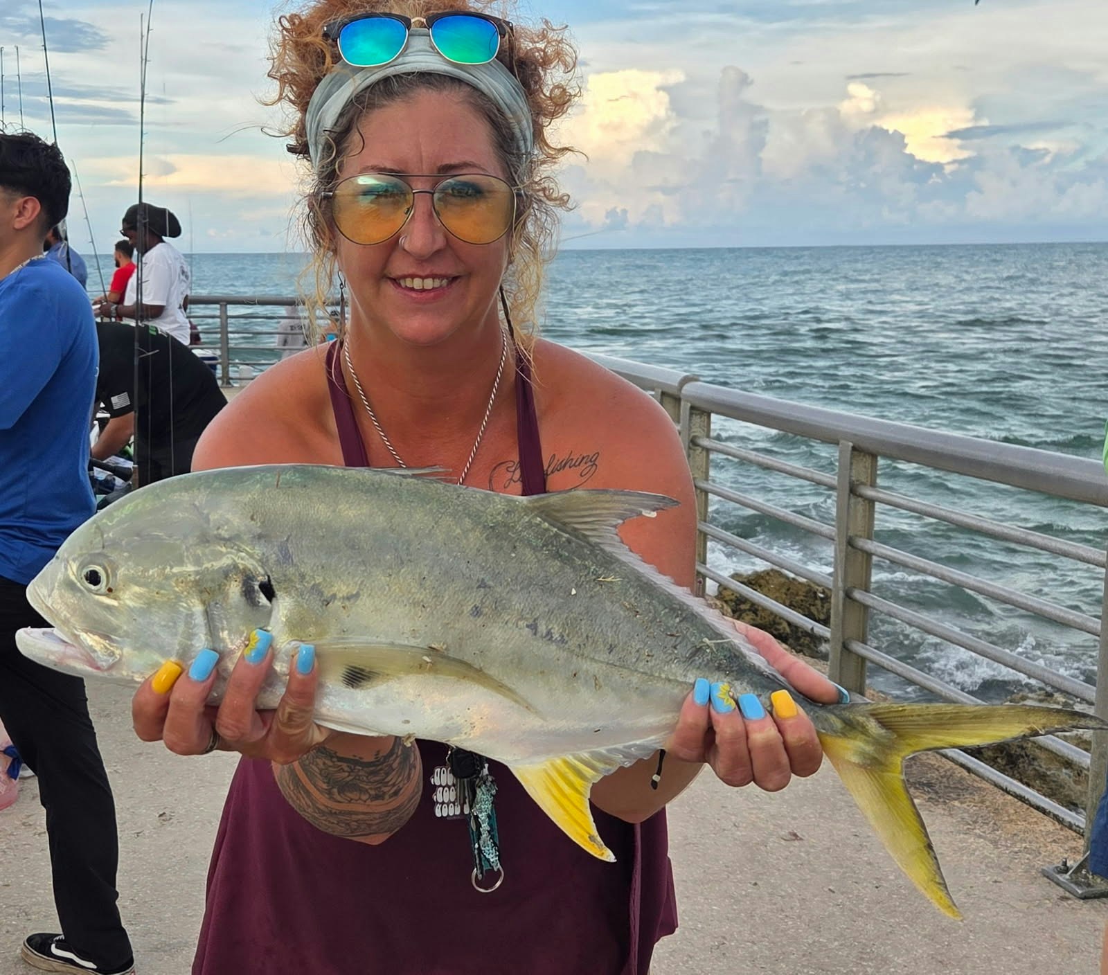 A person holding a large fish on a pier by the ocean, with a railing and other people in the background.