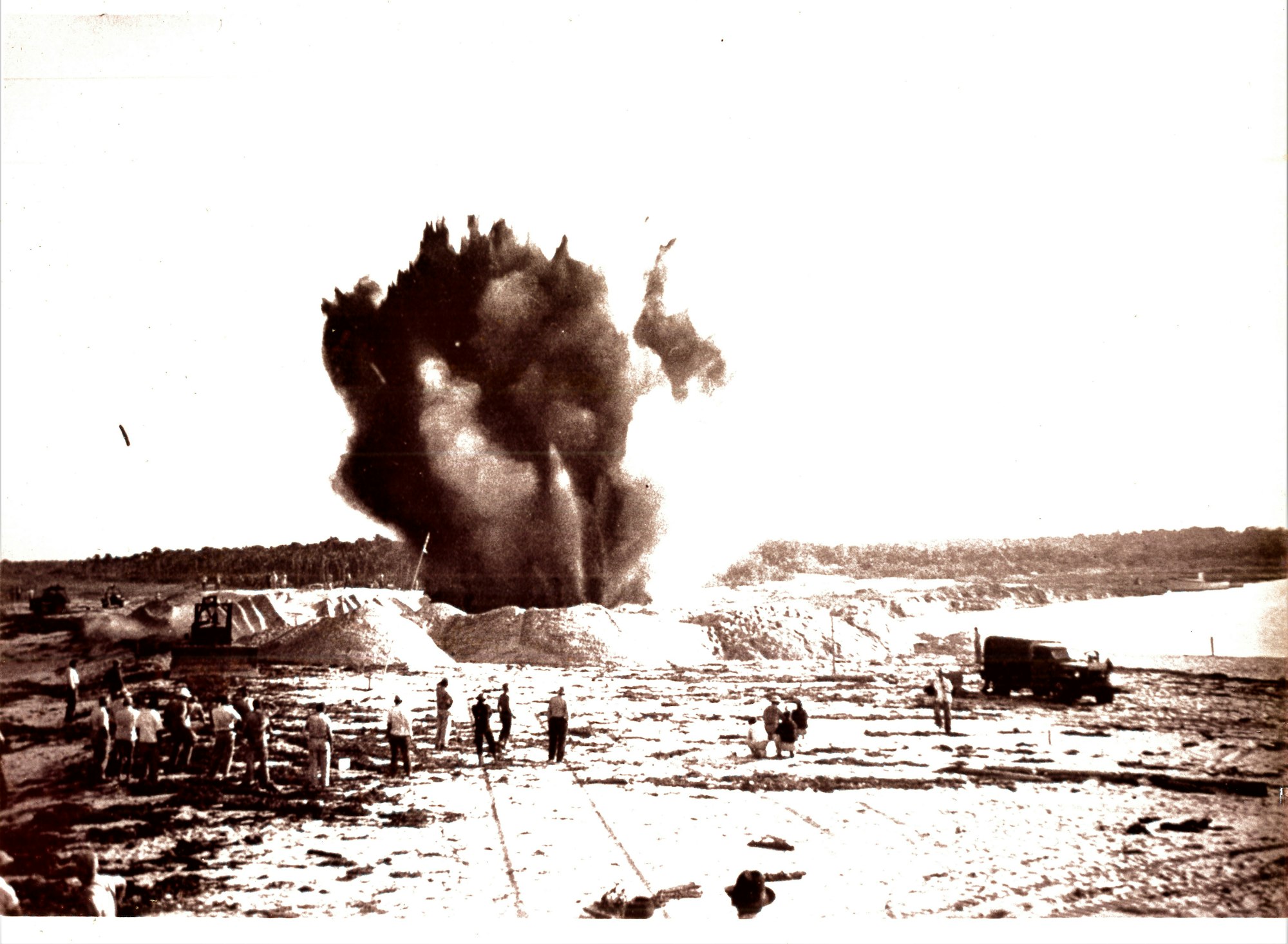 The image shows a large explosion with smoke rising, surrounded by people and machinery on a dirt landscape.