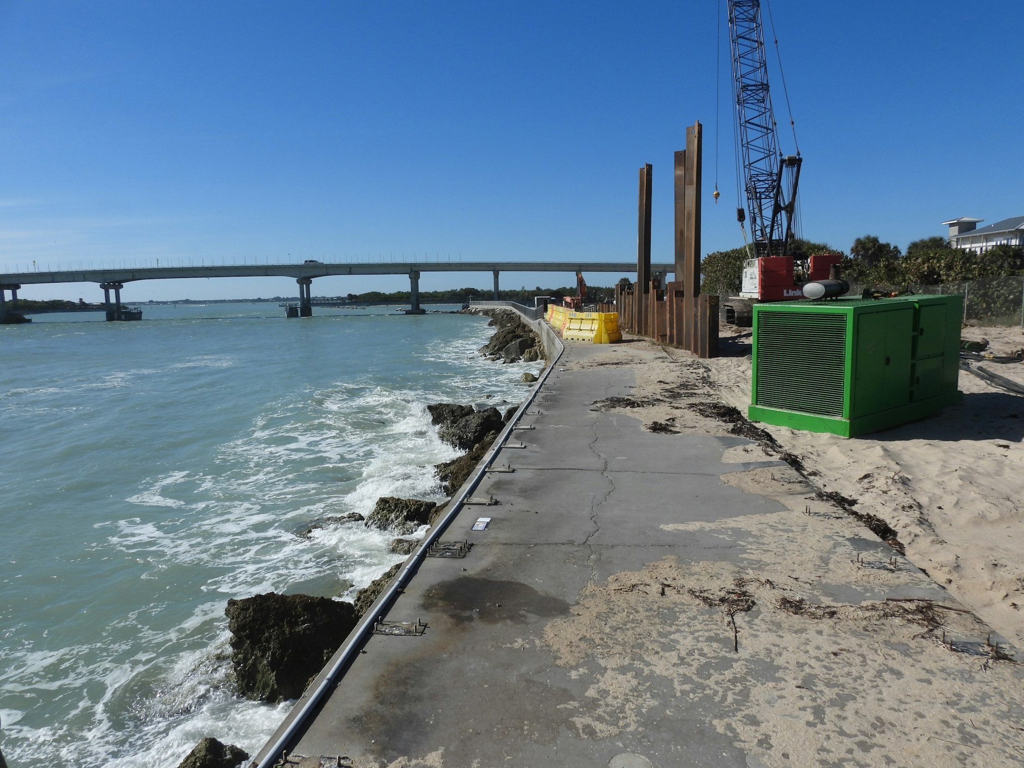 Coastal construction site with rocks, waves, a bridge, crane, and equipment under a clear blue sky.