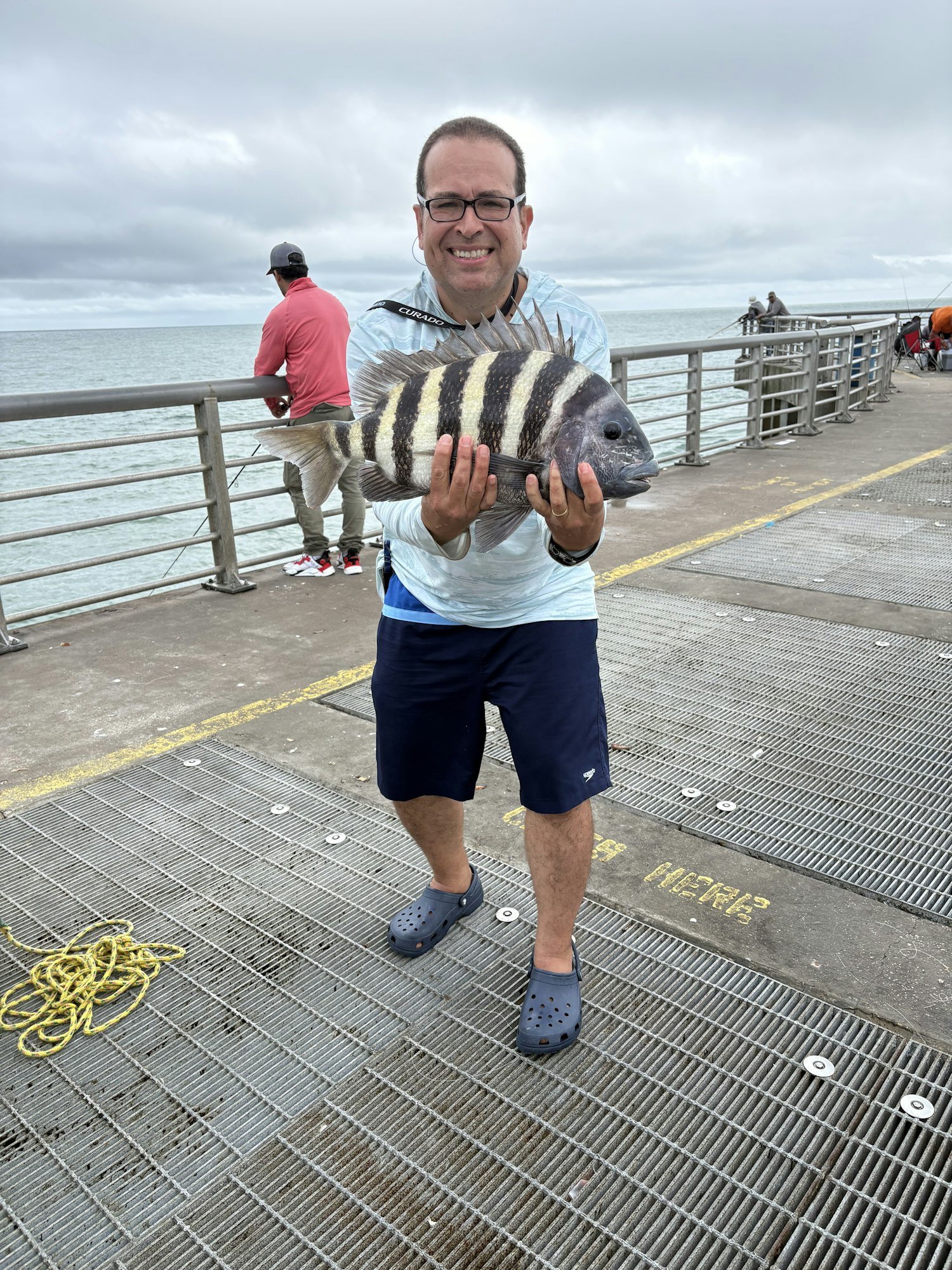 A person on a pier holds a striped fish, smiling, with ocean and fishing activities in the background.