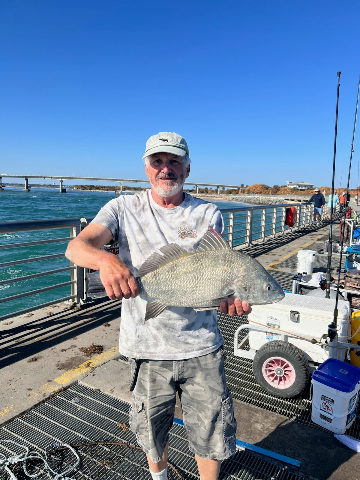 A man is holding a large fish on a fishing pier, with a clear blue sky and water in the background.