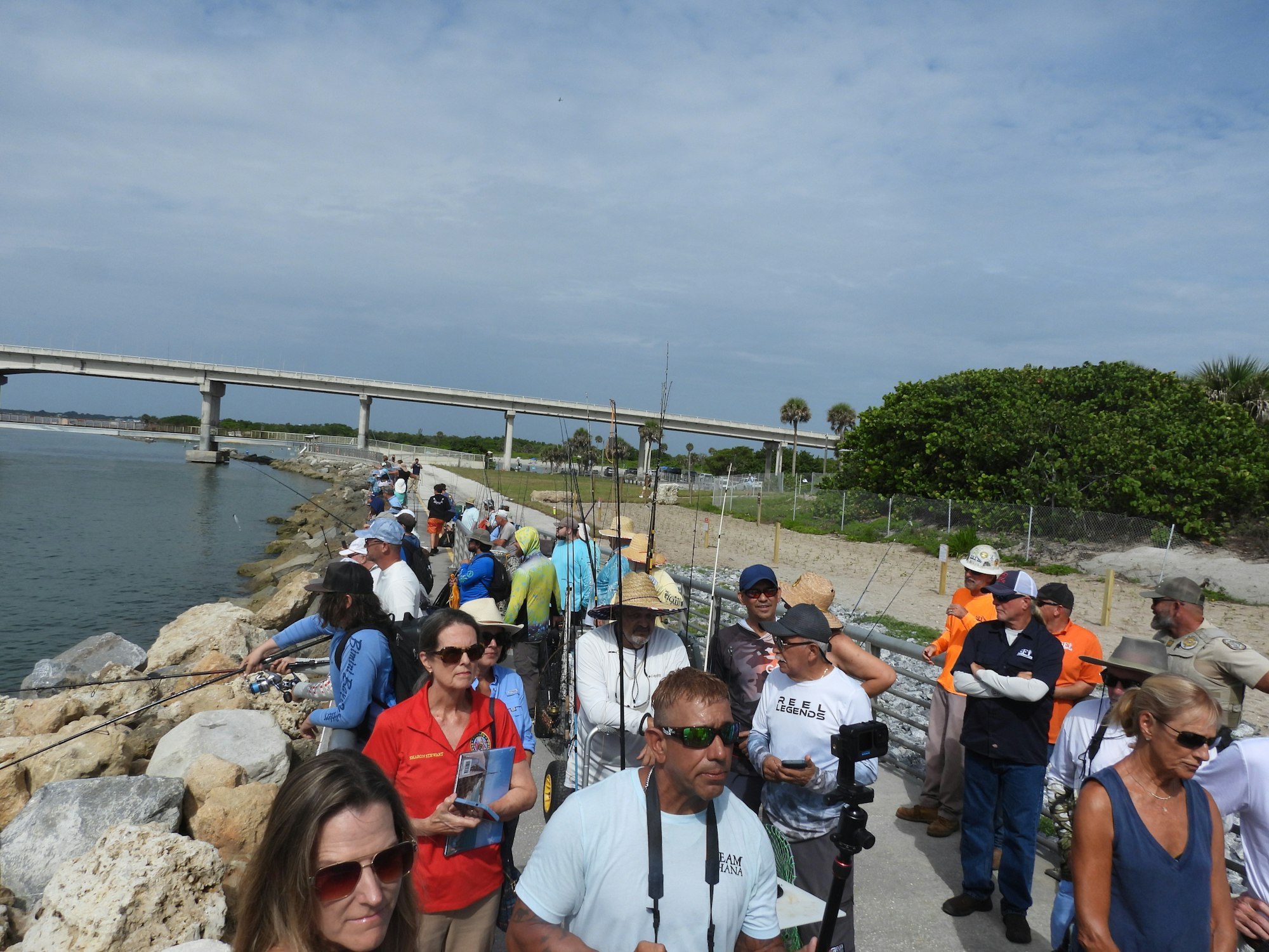 A group of people fishing along a rocky waterfront with a bridge in the background.