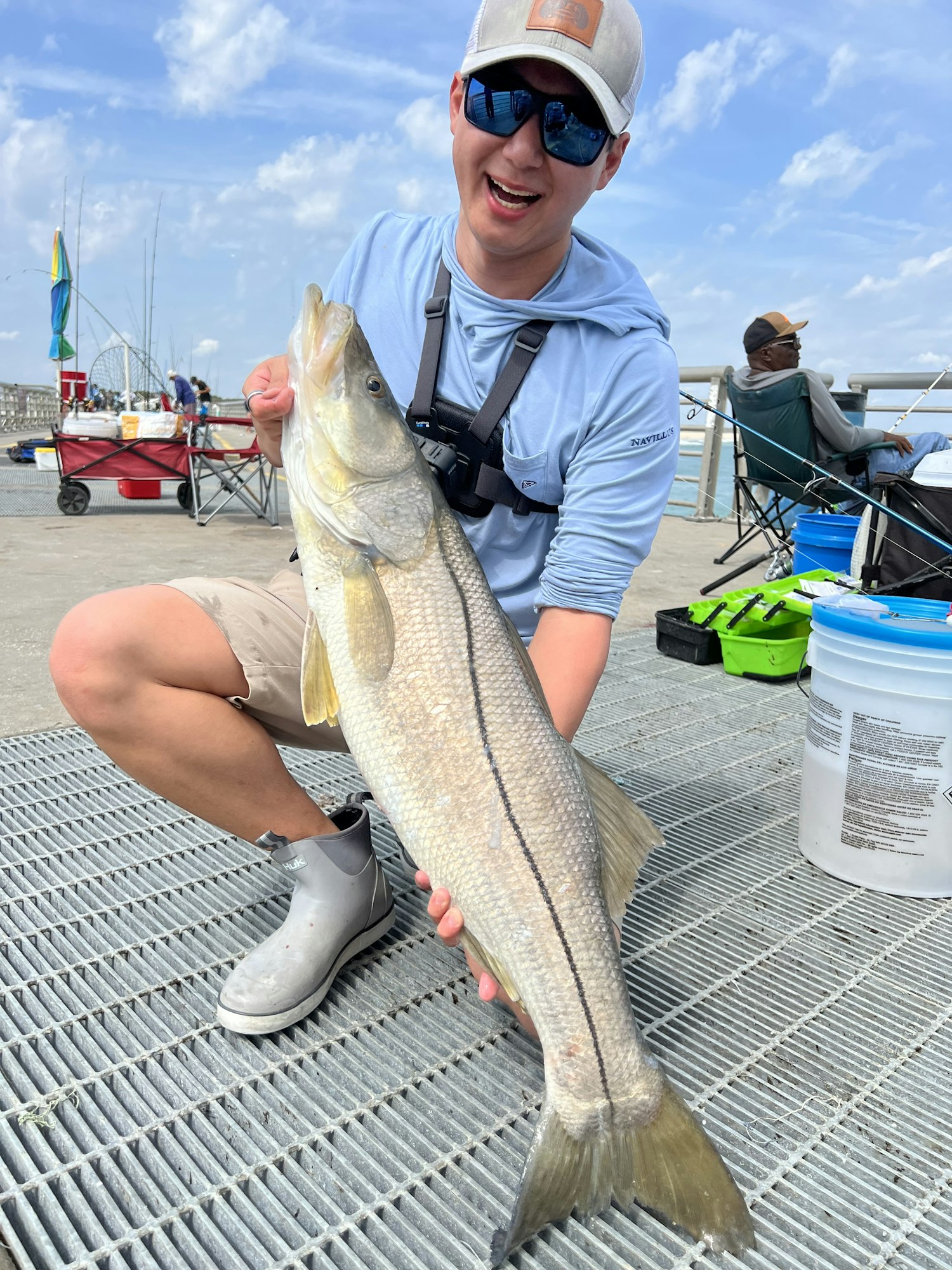 A man holding a large fish at a pier, with fishing gear and other people in the background.