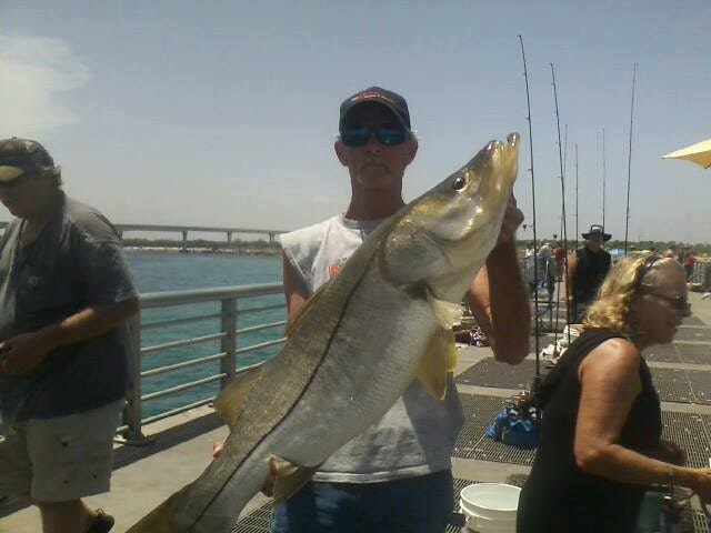 A man holds a large fish at a pier, with several fishing rods and other people in the background enjoying the day.