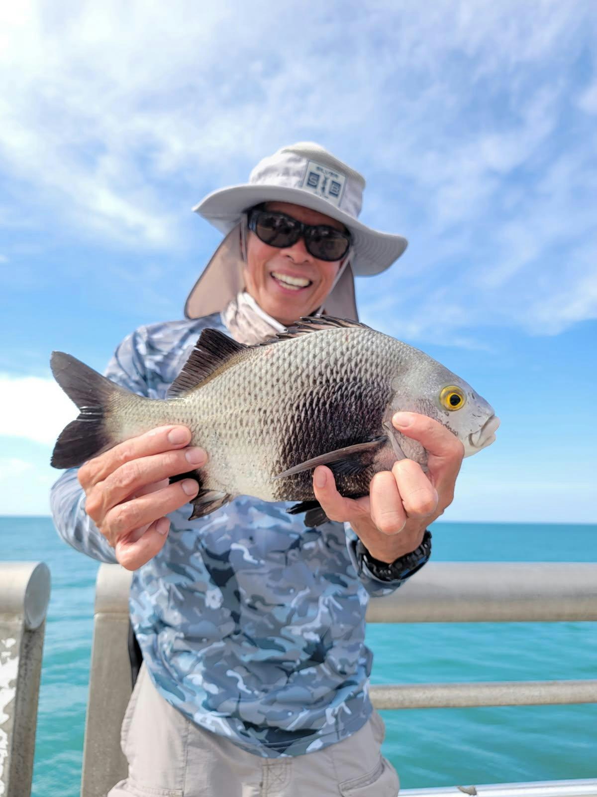 A person holding a fish, wearing a wide-brimmed hat and sunglasses, with the sea in the background.