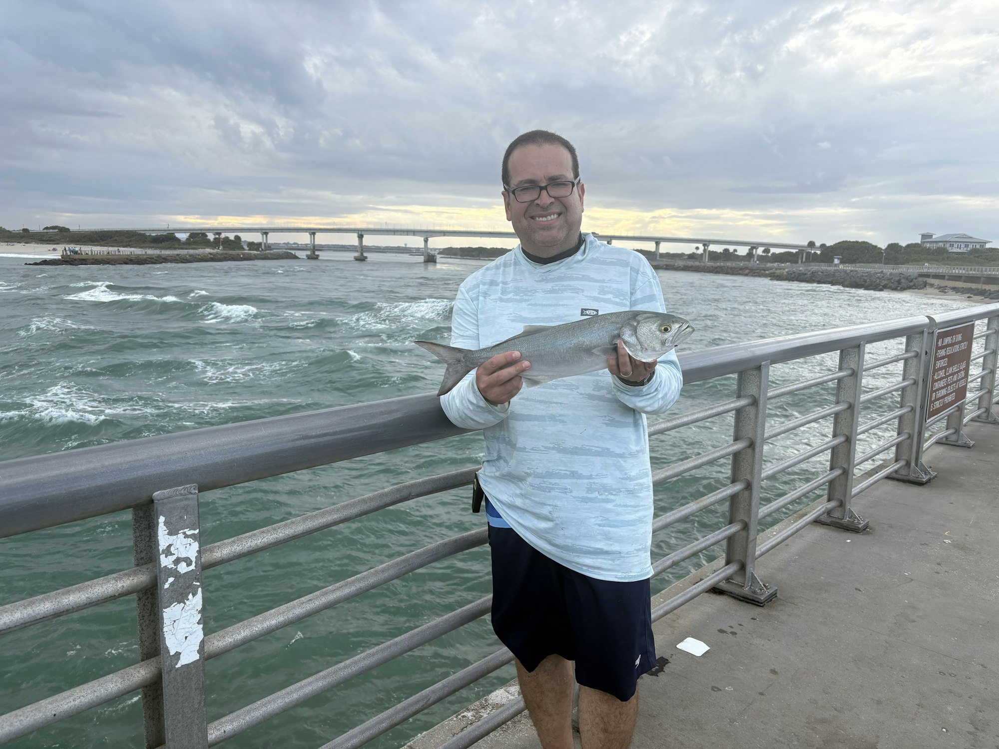 A person is standing on a pier, proudly holding a fish, with waves and a bridge visible in the background under a cloudy sky.