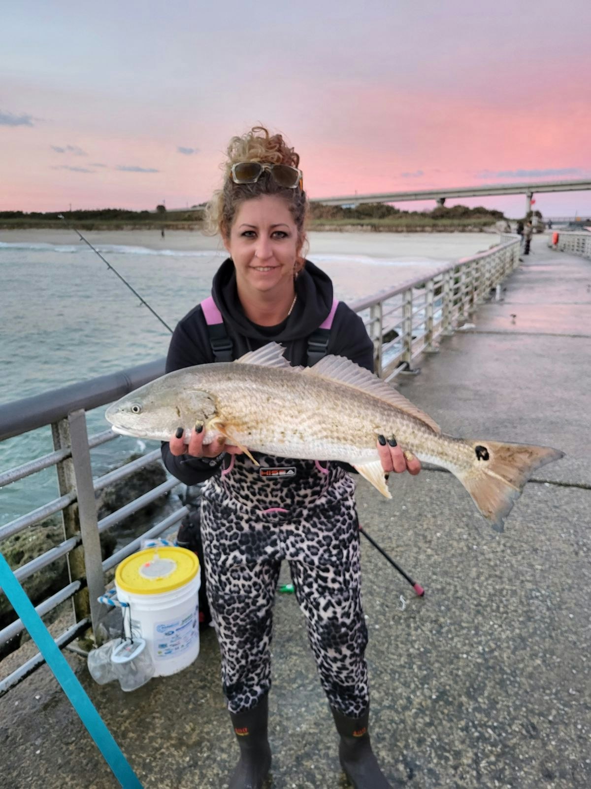 A woman on a pier proudly holds a large fish against a colorful sunset backdrop, wearing stylish fishing gear.