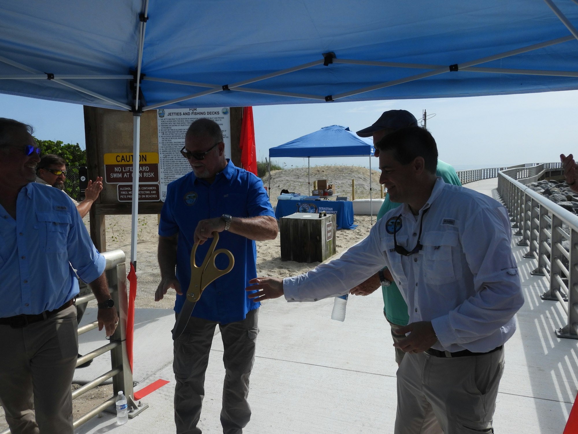 A group of people under a canopy, one holding large scissors, likely at a ribbon-cutting event near a beach.
