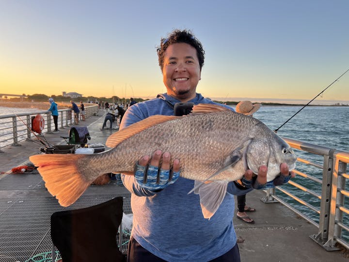 A smiling person holds a large fish on a pier at sunset, with other anglers visible in the background.