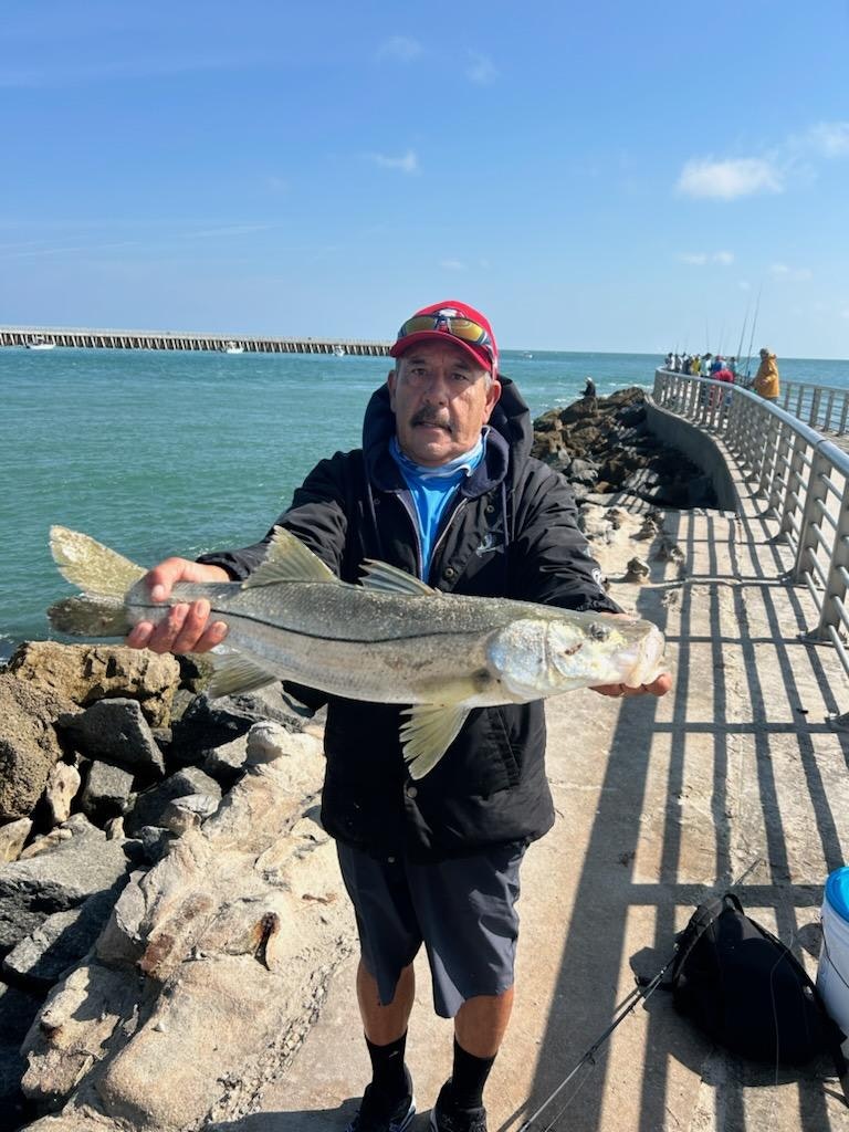 A person holding a large fish on a pier by the sea, with fishing rods and a bucket nearby.