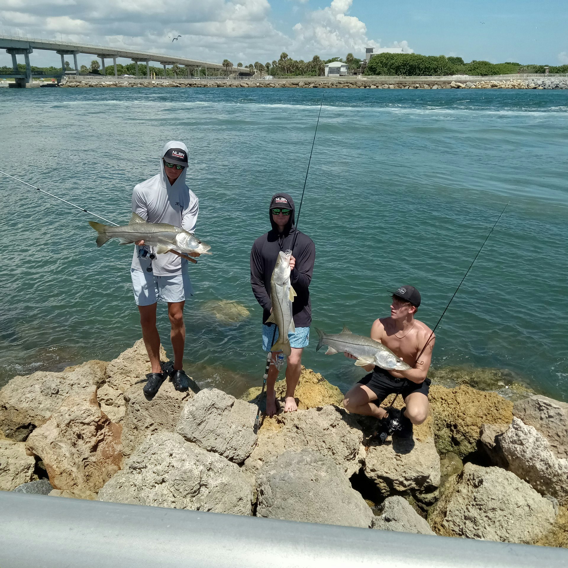 Three people holding fish near the water on rocks; fishing rods visible.