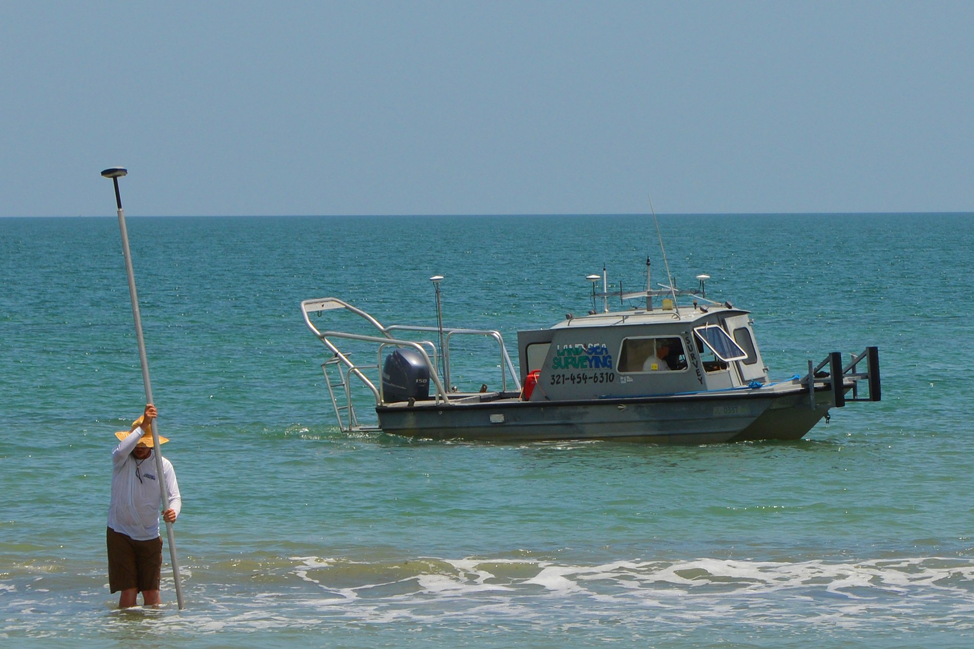 man standing in surf with equipment with boat in background