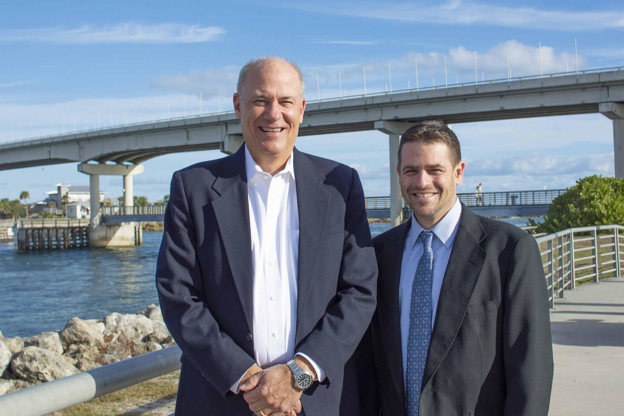 two men standing in front of bridge over inlet in background