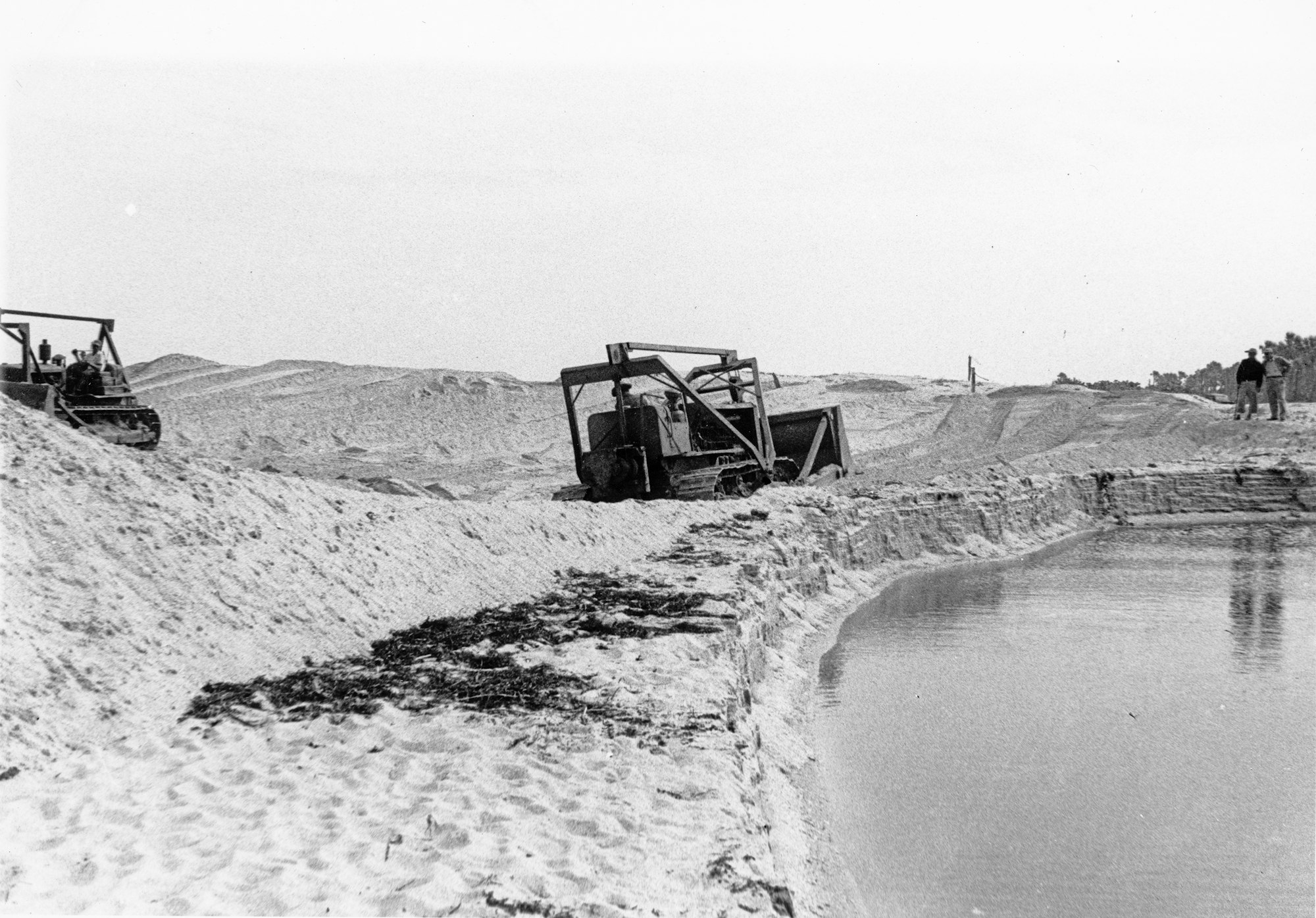 An industrial site featuring machinery on sandy terrain, near a body of water, with workers visible in the background.