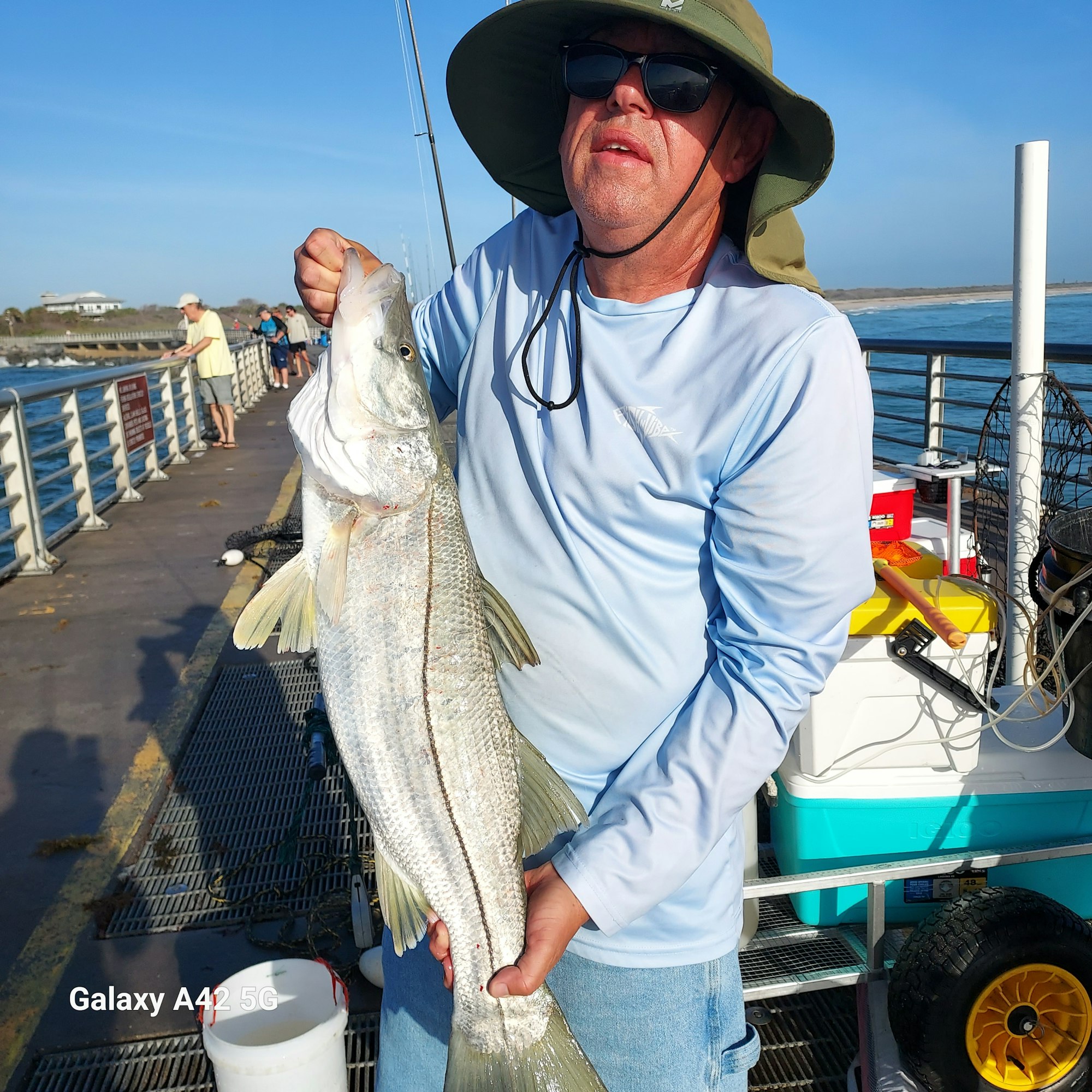 A man holding a large fish on a pier, wearing sunglasses and a hat, with water and fishing gear in the background.
