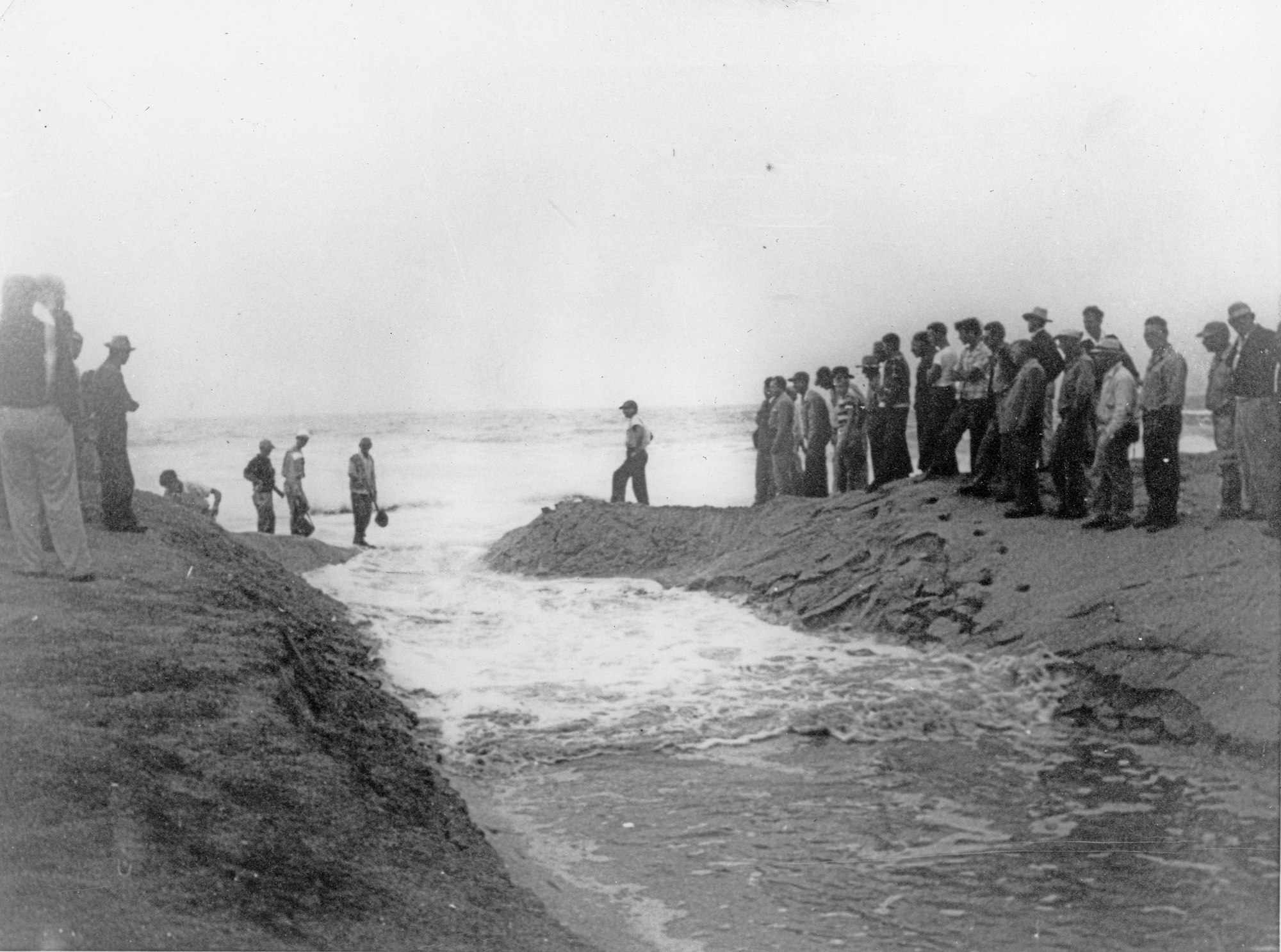 A group of people watches a scene by the shore, with some individuals near the water and others standing on the sand.