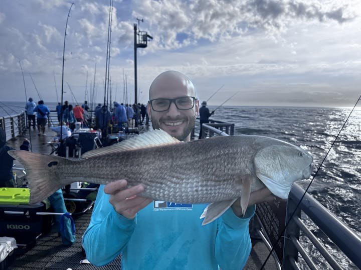 A person holds a sizable fish, smiling on a fishing pier with others in the background and a scenic ocean view.