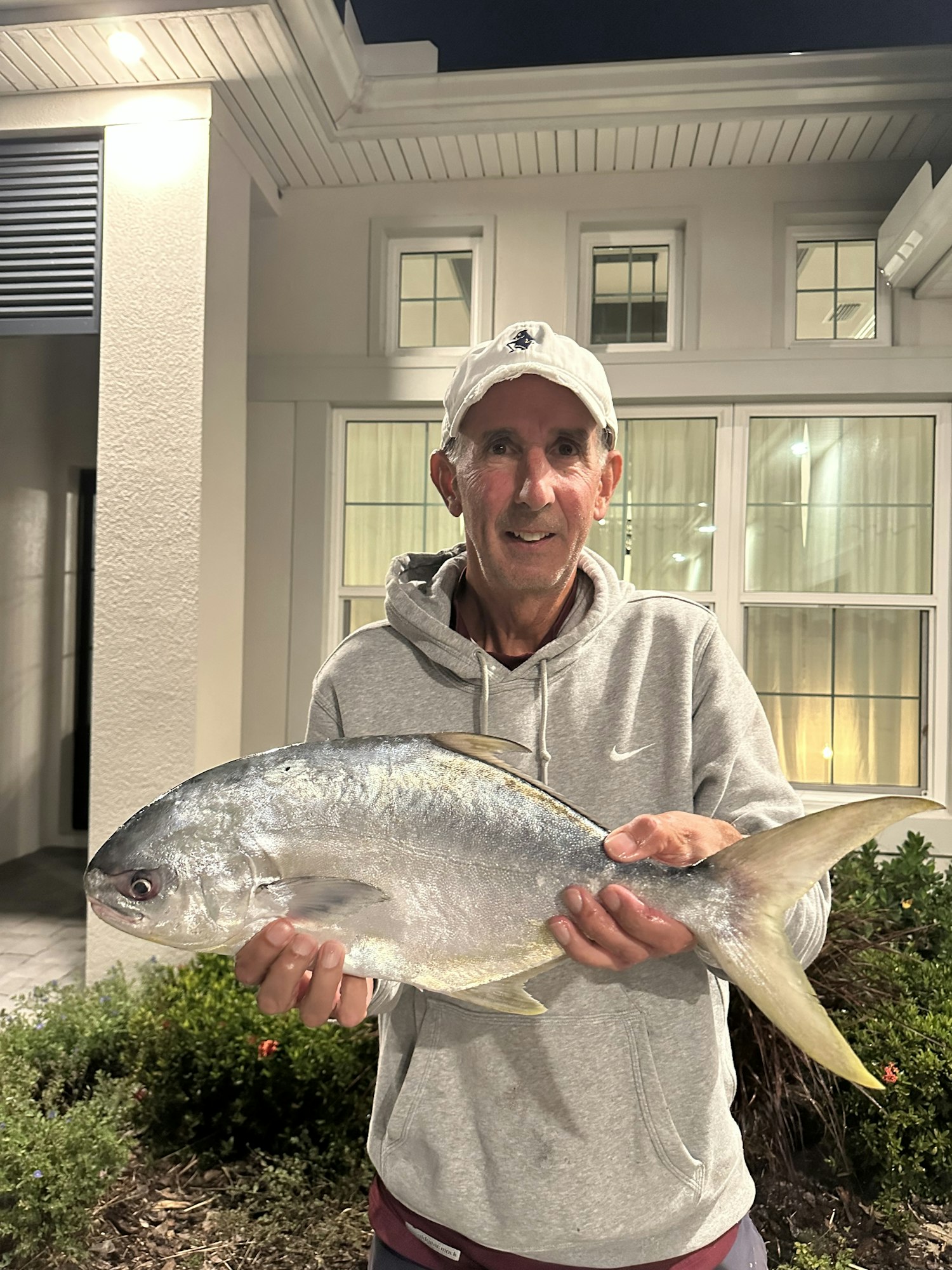A man is holding a large fish outside a house at night, smiling and dressed in casual clothing.