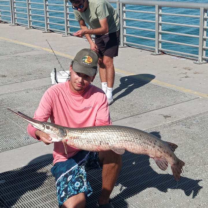 A person on a pier holds a large fish while another person is fishing in the background, enjoying a day outdoors.